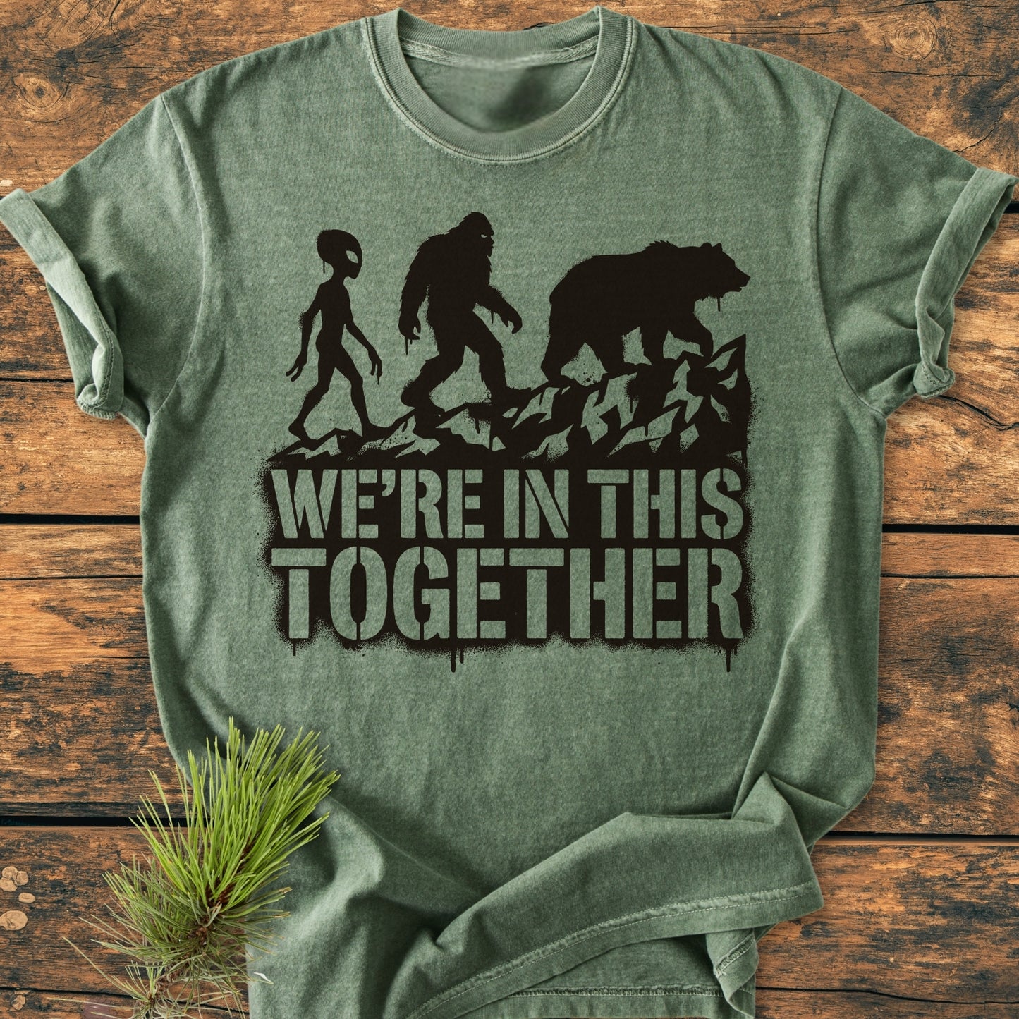 In This Together - Vintage Wash Tee features black silhouettes of an alien, Bigfoot, and a bear over a rocky ridge, with the slogan WE'RE IN THIS TOGETHER on soft green fabric.