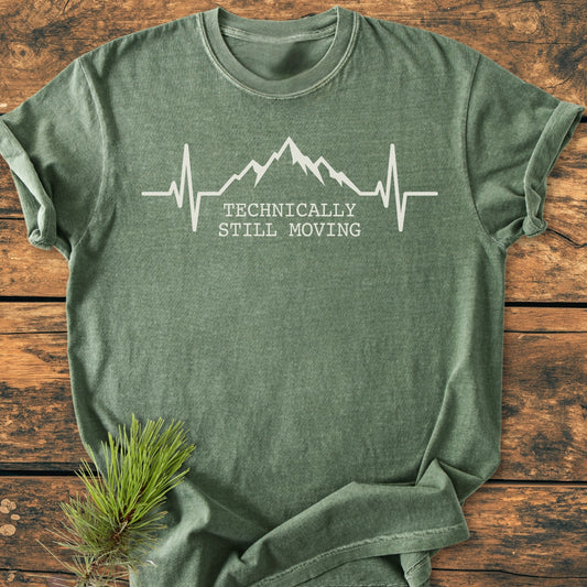 The Still Moving - Vintage Wash Tee features a heather-green color with a heartbeat and mountain graphic, and the phrase TECHNICALLY STILL MOVING in white on a soft, relaxed-fit cotton shirt.