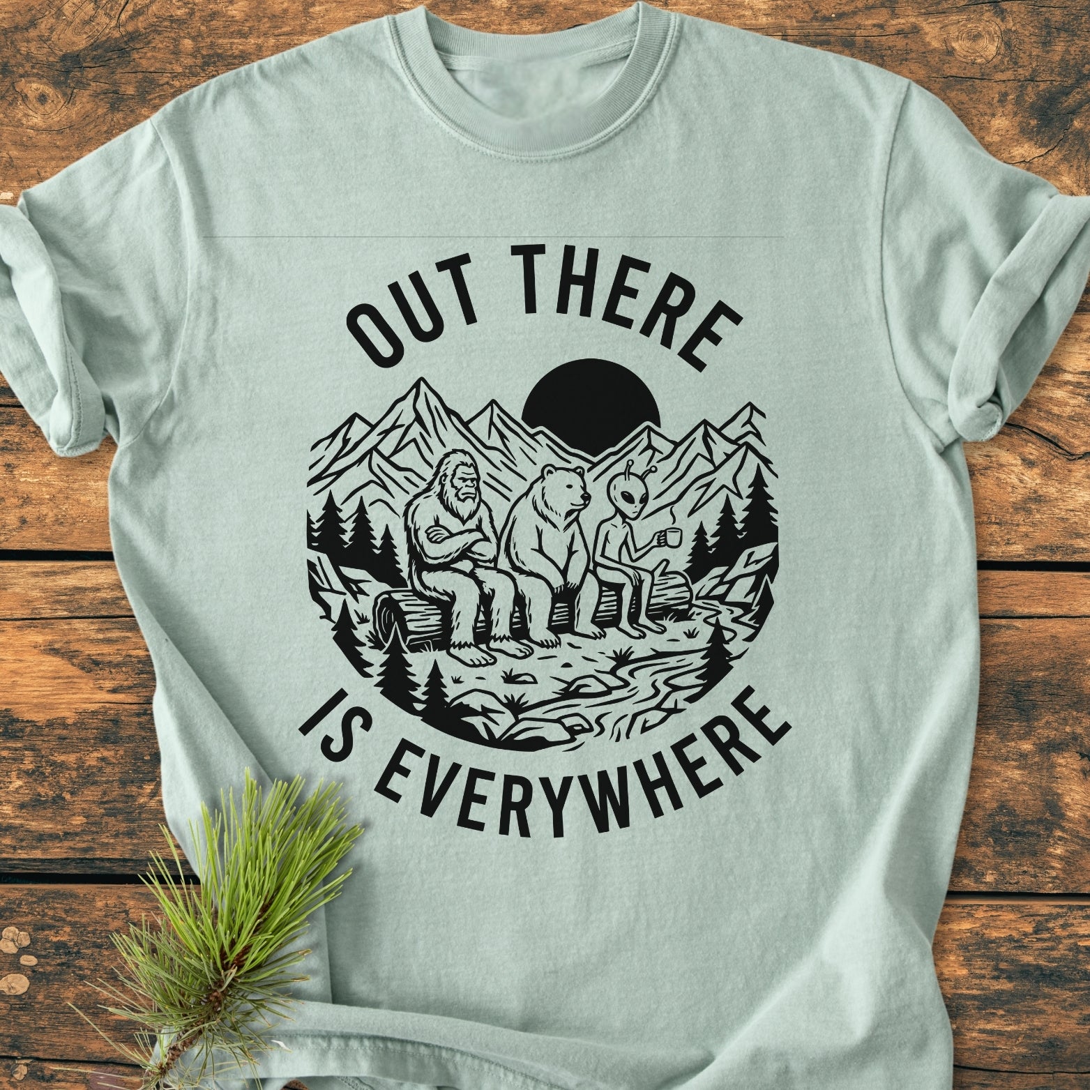 Out There Is Everywhere - Vintage Wash Tee features a graphic of Bigfoot, a bear, and an alien on a log with the slogan, showcasing its relaxed fit and soft garment-dyed fabric.