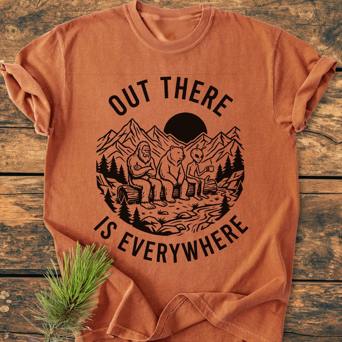Out There Is Everywhere - Vintage Wash Tee: Features a wilderness scene with Bigfoot, bear, and alien holding mugs, set against a rustic pine sprig, highlighting its relaxed fit and cozy fabric.