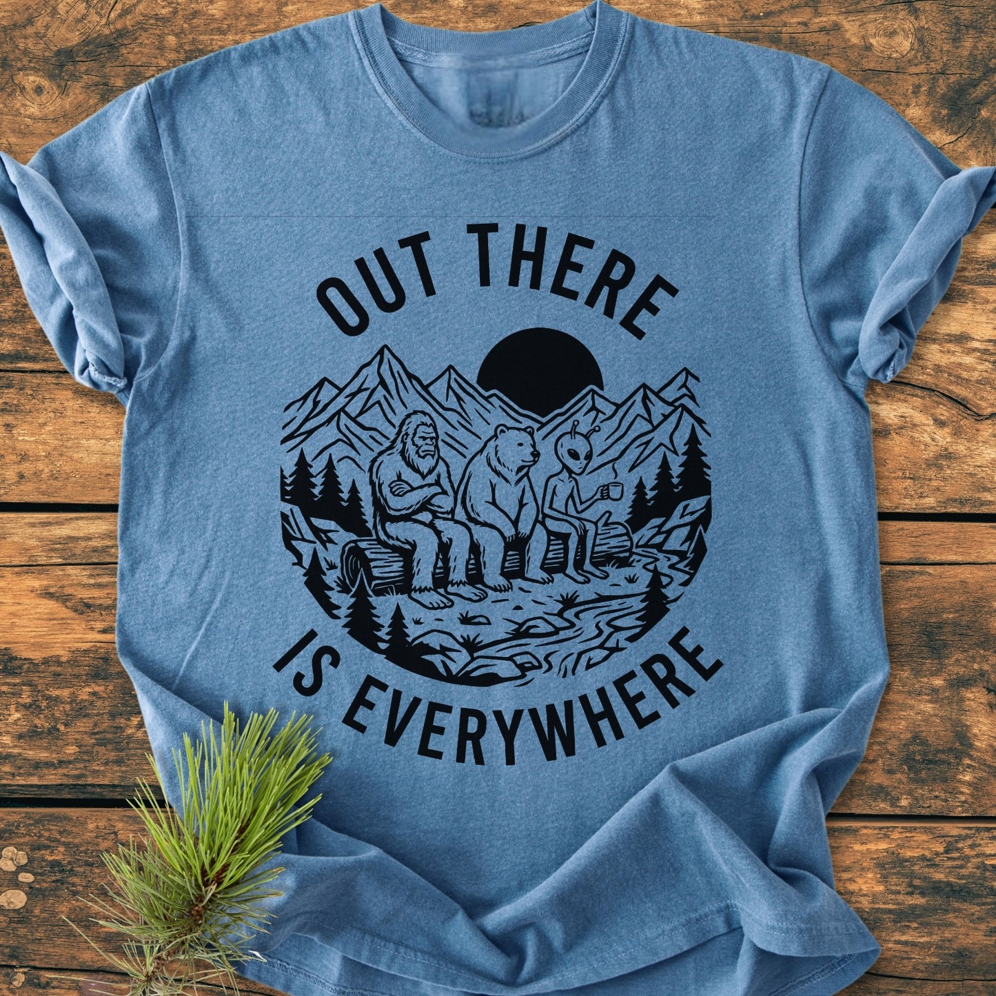 Out There Is Everywhere - Vintage Wash Tee features a mountain scene with Bigfoot, bear, and alien illustration on a heather-blue backdrop, styled with a pine sprig for rustic appeal.