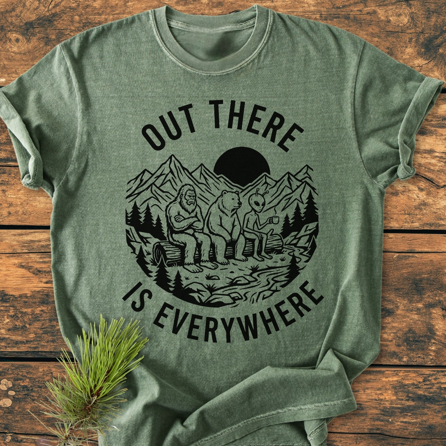 Out There Is Everywhere - Vintage Wash Tee featuring a wilderness scene with Bigfoot, bear, and alien. Laid flat, showcasing its relaxed fit and durable stitching on a rustic wooden surface.