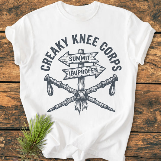 Creaky Knee Corps - Vintage Wash Tee featuring a playful hiking-themed design with CREAKY KNEE CORPS text, a signpost, and crossed hiking poles, laid on a wood-grain surface with a pine branch.