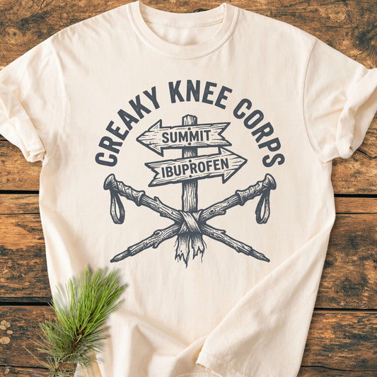 Creaky Knee Corps - Vintage Wash Tee featuring a humorous logo with a rustic signpost and crossed hiking poles, symbolizing the hiking spirit. Laid on a wood surface, highlighting its relaxed fit and durability.