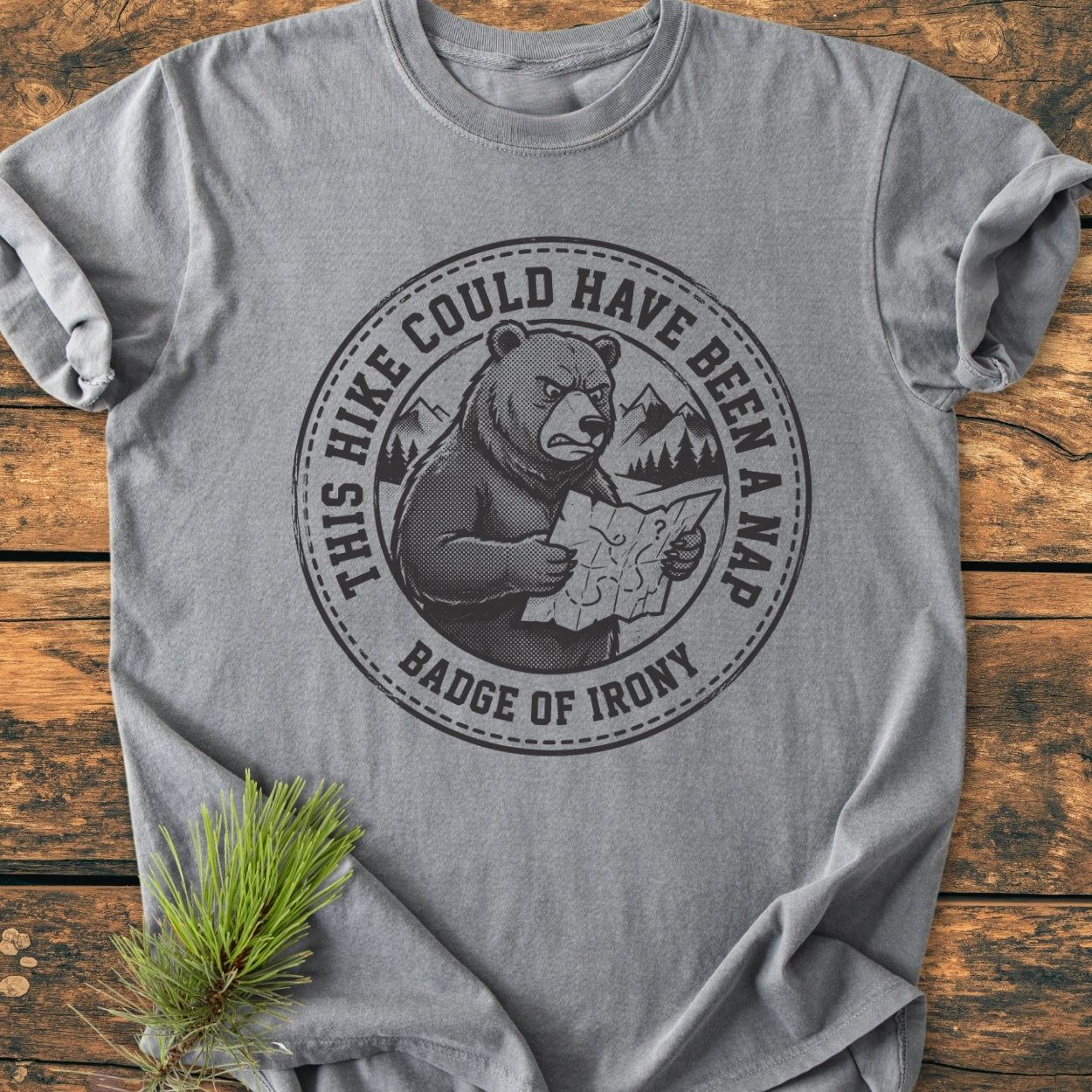 Could Have Been A Nap - Vintage Wash Tee featuring an irritated bear graphic with mountains, emphasizing its relaxed fit and soft-washed fabric. Laid on wood with pine sprig accent.