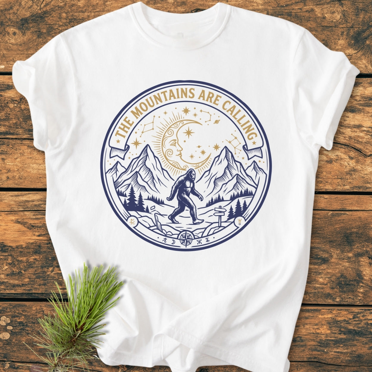 The Mountains Are Calling - Vintage Wash Tee features a two-tone outdoor graphic with mountains, pine trees, and Bigfoot, laid on wood beside a pine sprig, showcasing its adventurous design.