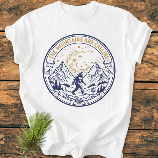 The Mountains Are Calling - Vintage Wash Tee features a two-tone outdoor graphic with mountains, pine trees, and Bigfoot, laid on wood beside a pine sprig, showcasing its adventurous design.