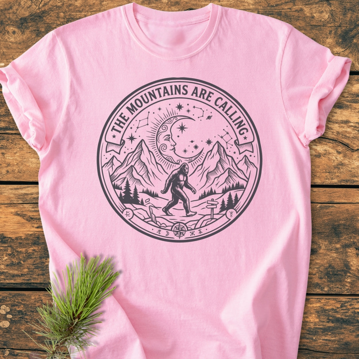 The Mountains Are Calling - Vintage Wash Tee featuring Bigfoot, pine trees, and mountain scenery within a circular motif on a light-pink T-shirt, emphasizing a mystical, outdoorsy theme.
