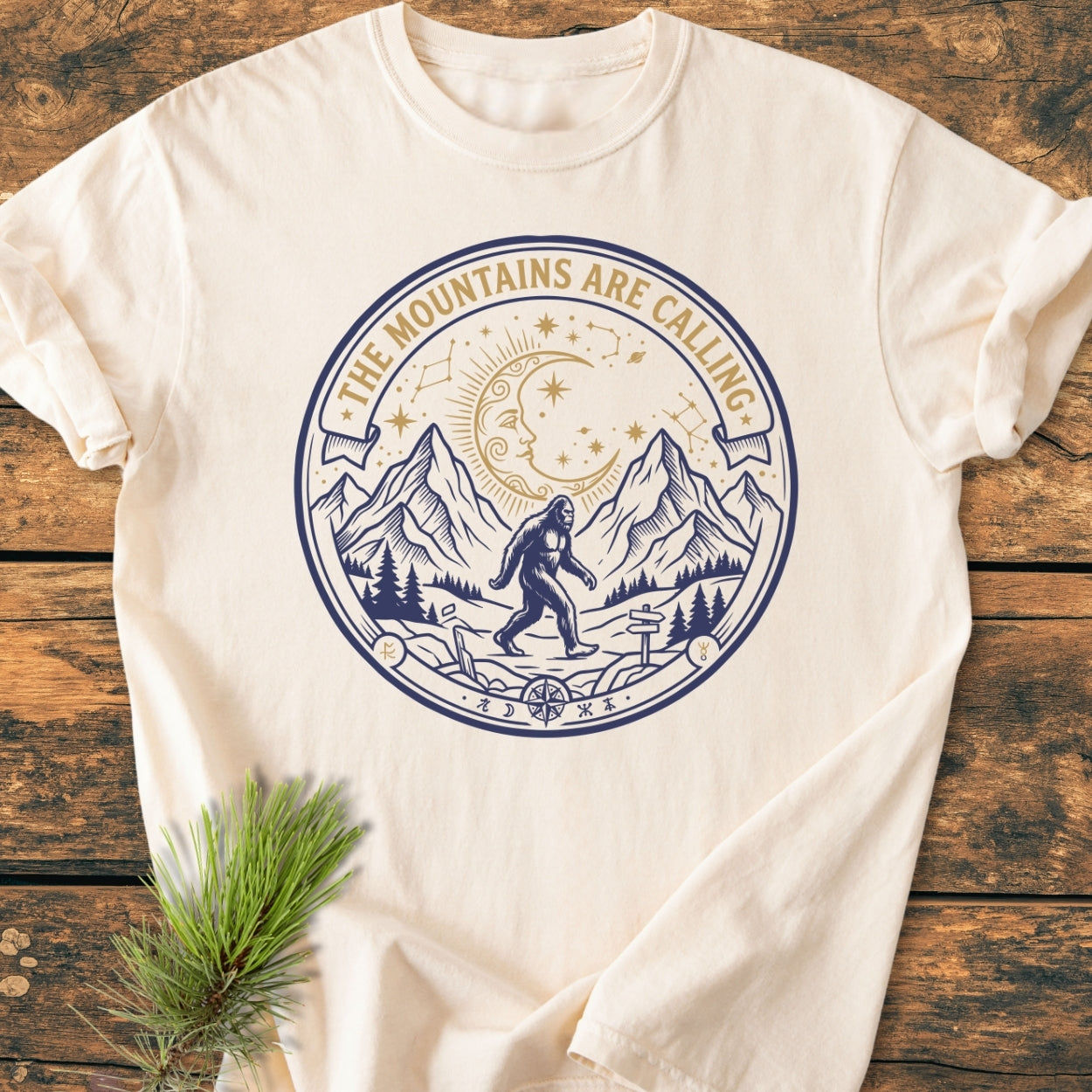 The Mountains Are Calling - Vintage Wash Tee features a two-tone graphic of Sasquatch, mountains, and celestial elements, laid on a rustic surface with a pine sprig beside it.