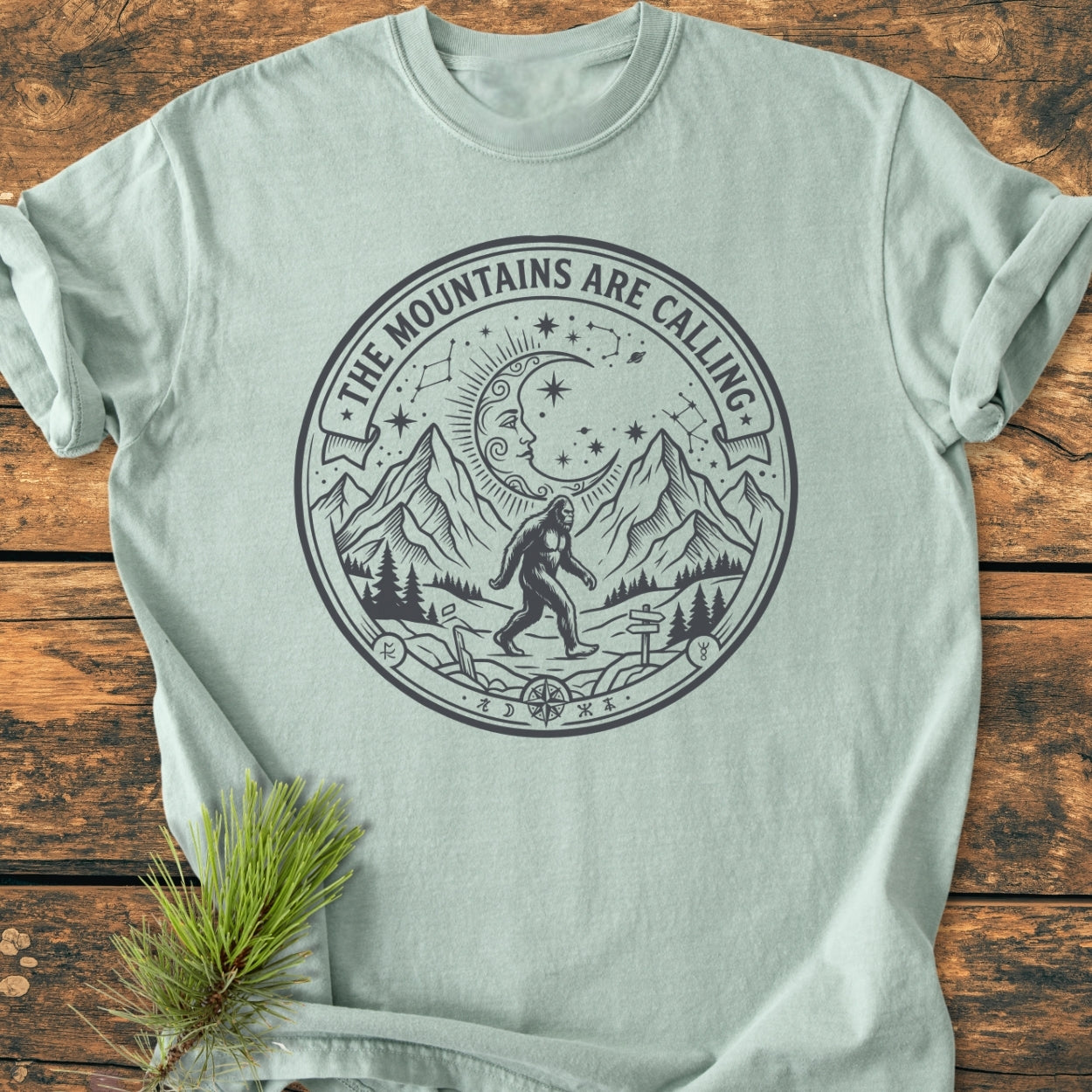 The Mountains Are Calling - Vintage Wash Tee features a circular mountain and Bigfoot graphic, laid flat on wood, showcasing Comfort Colors' soft, relaxed fit, and durable garment-dyed cotton.