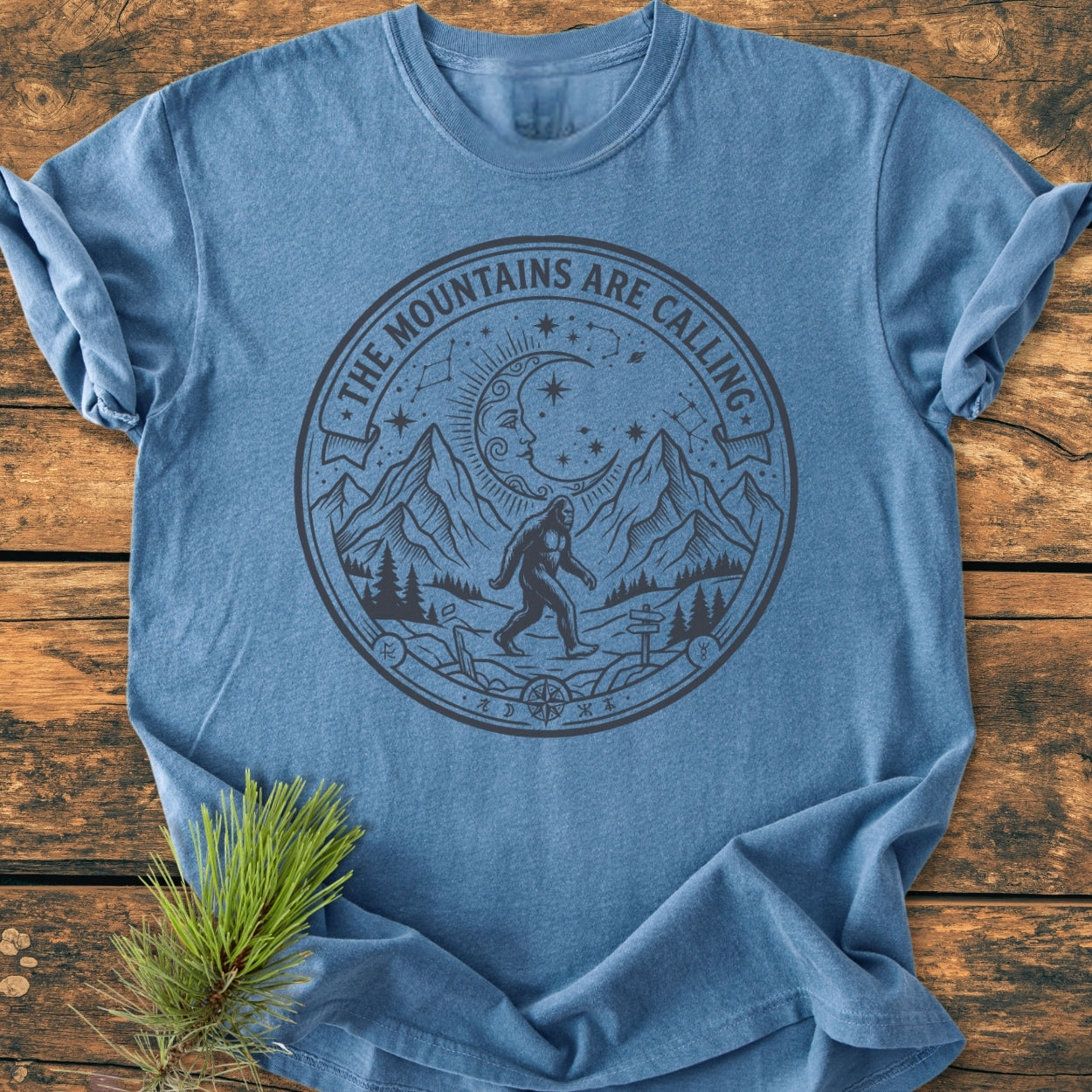The Mountains Are Calling - Vintage Wash Tee features a rugged mountain scene with Sasquatch, moon, and stars in a circular graphic, laid flat on a wooden surface with a pine sprig.
