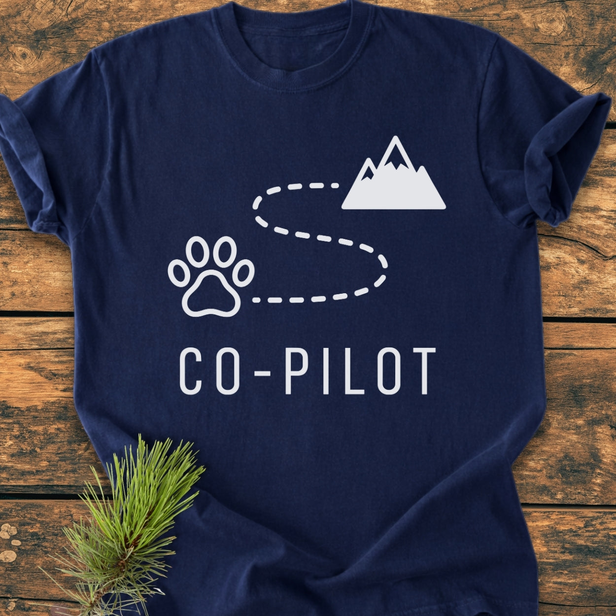 Co-Pilot Dog - Vintage Wash Tee features a playful design with a paw print, winding trail, and mountain silhouette, symbolizing a hiking adventure with your pet companion. Laid on a wooden surface.