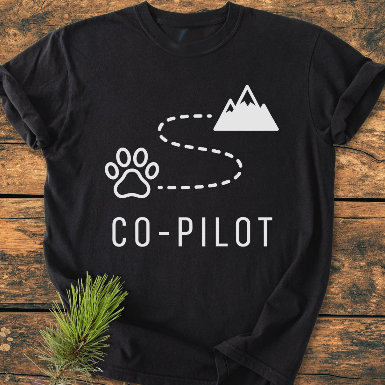 Co-Pilot Dog - Vintage Wash Tee features a playful design with a paw print, path, and mountain silhouette on a black T-shirt, laid on wood with a pine twig.