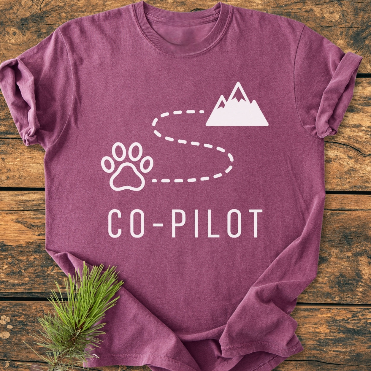 Heather-purple Co-Pilot Dog - Vintage Wash Tee featuring a dog-paw icon, dashed line, and mountain silhouette design. Laid flat on wood, with a pine sprig accentuating its cozy, relaxed style.