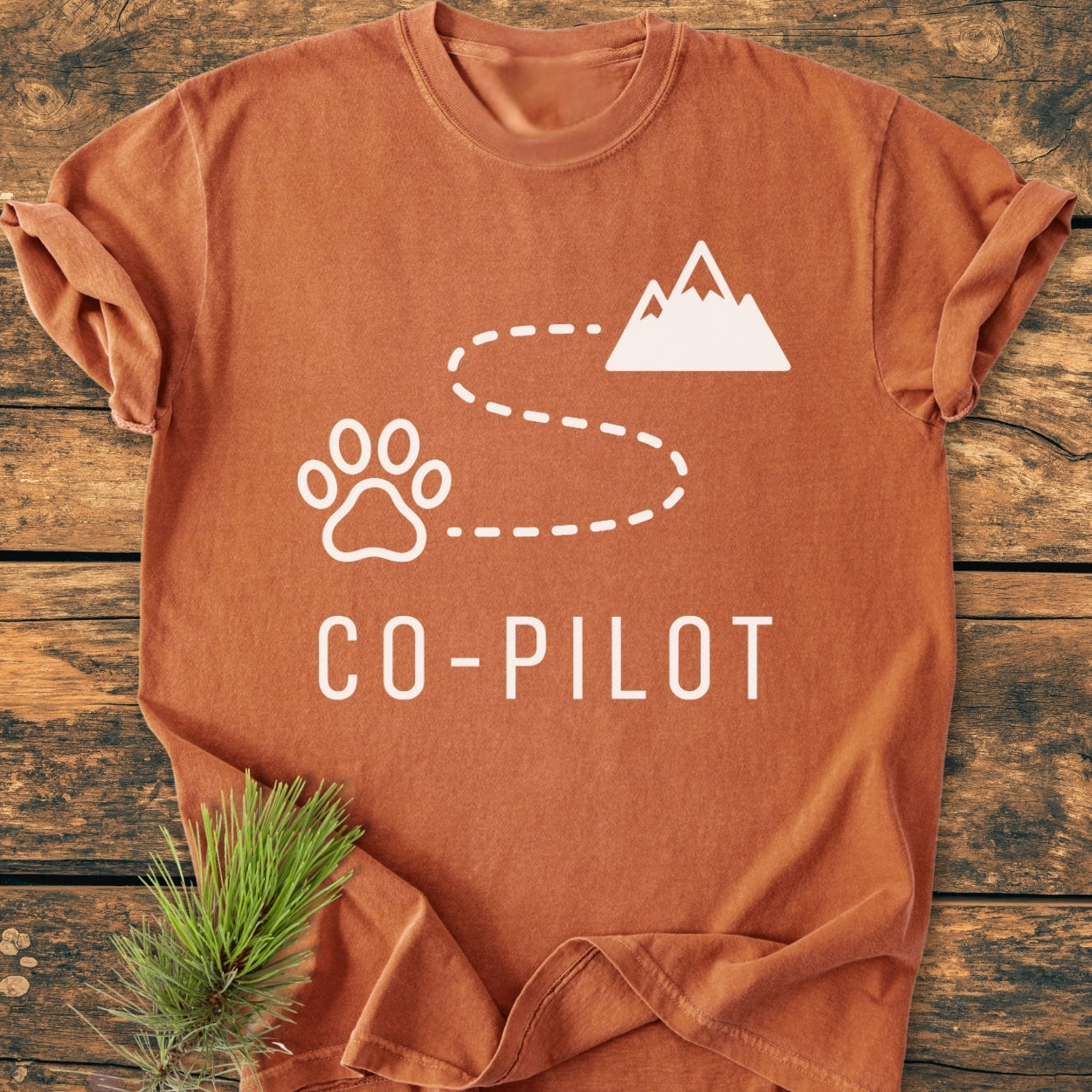 Co-Pilot Dog - Vintage Wash Tee: Rust-orange short-sleeve T-shirt featuring a white paw print and mountain graphic with CO-PILOT text, laid flat with a pine sprig for an outdoorsy touch.
