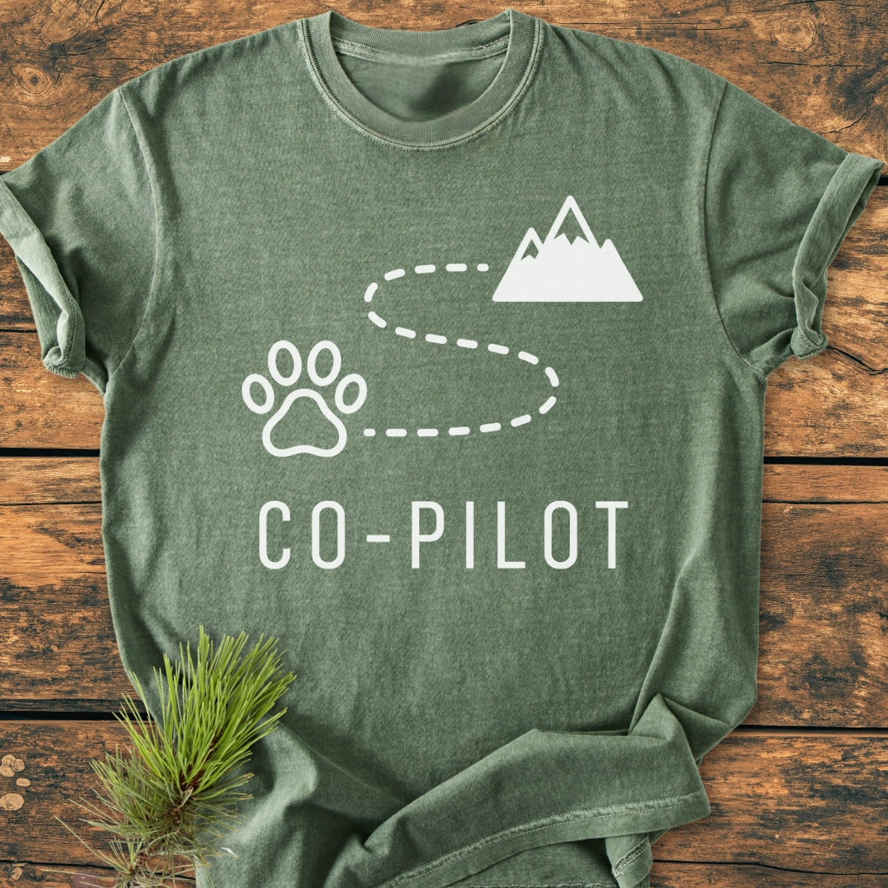 Co-Pilot Dog - Vintage Wash Tee in heather-green, featuring a white paw print, trail, and mountain graphic, laid on wood. Includes sprig of pine for rustic accent, highlighting relaxed fit and durability.
