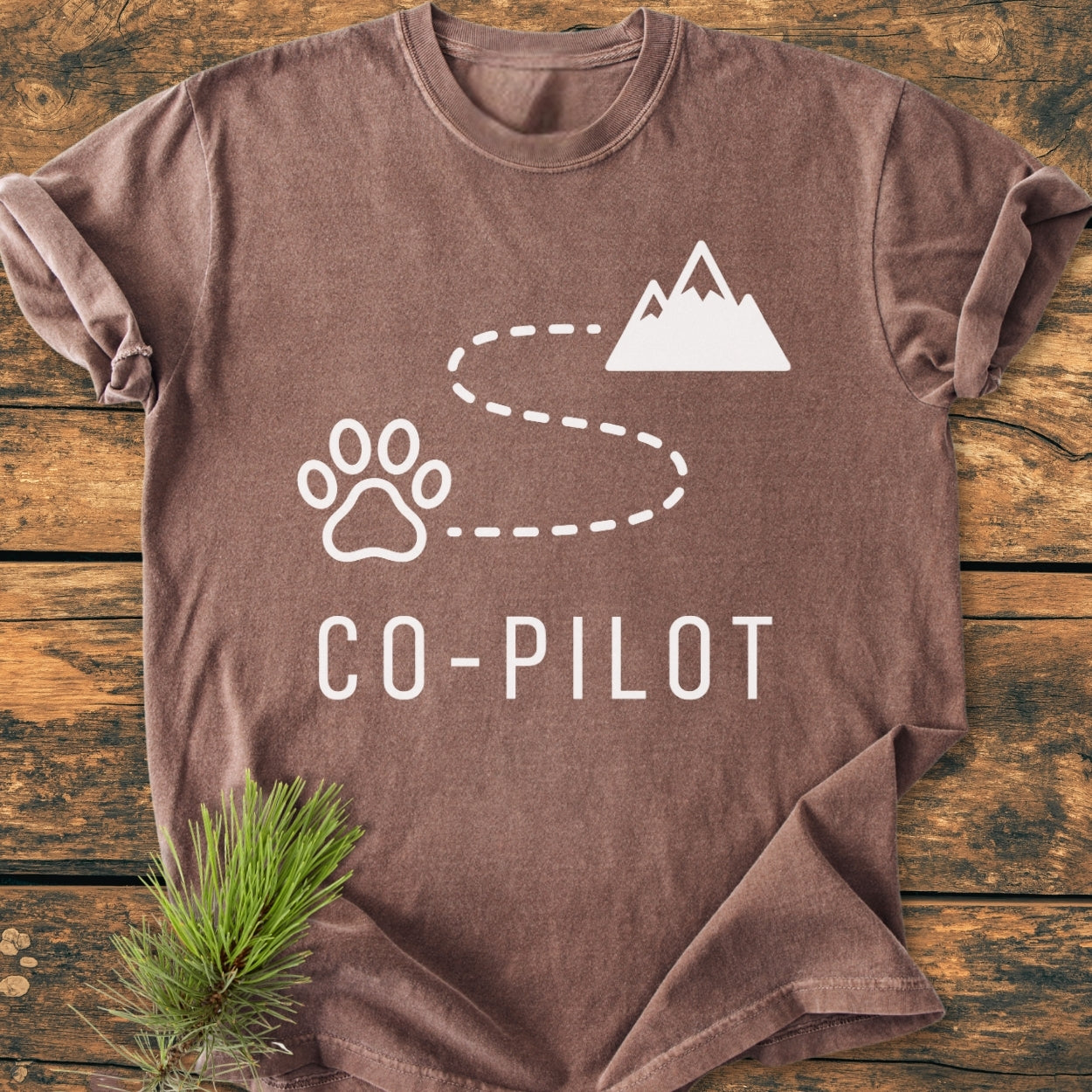 Co-Pilot Dog - Vintage Wash Tee featuring a white paw print, mountain peak, and trail graphic on a brown short-sleeved T-shirt, laid flat on a wooden table with a pine sprig nearby.