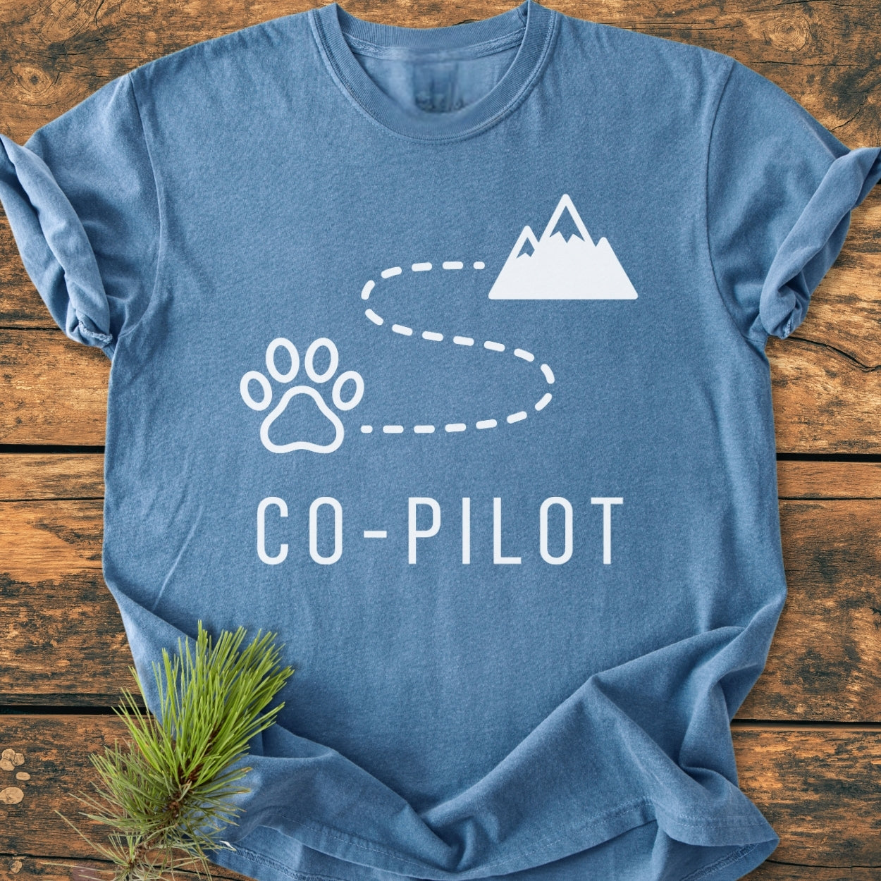 Co-Pilot Dog - Vintage Wash Tee features a paw print, trail, and mountain graphics in white on a blue background, with a pine sprig accent. Laid flat, showcasing its relaxed fit and durable stitching.
