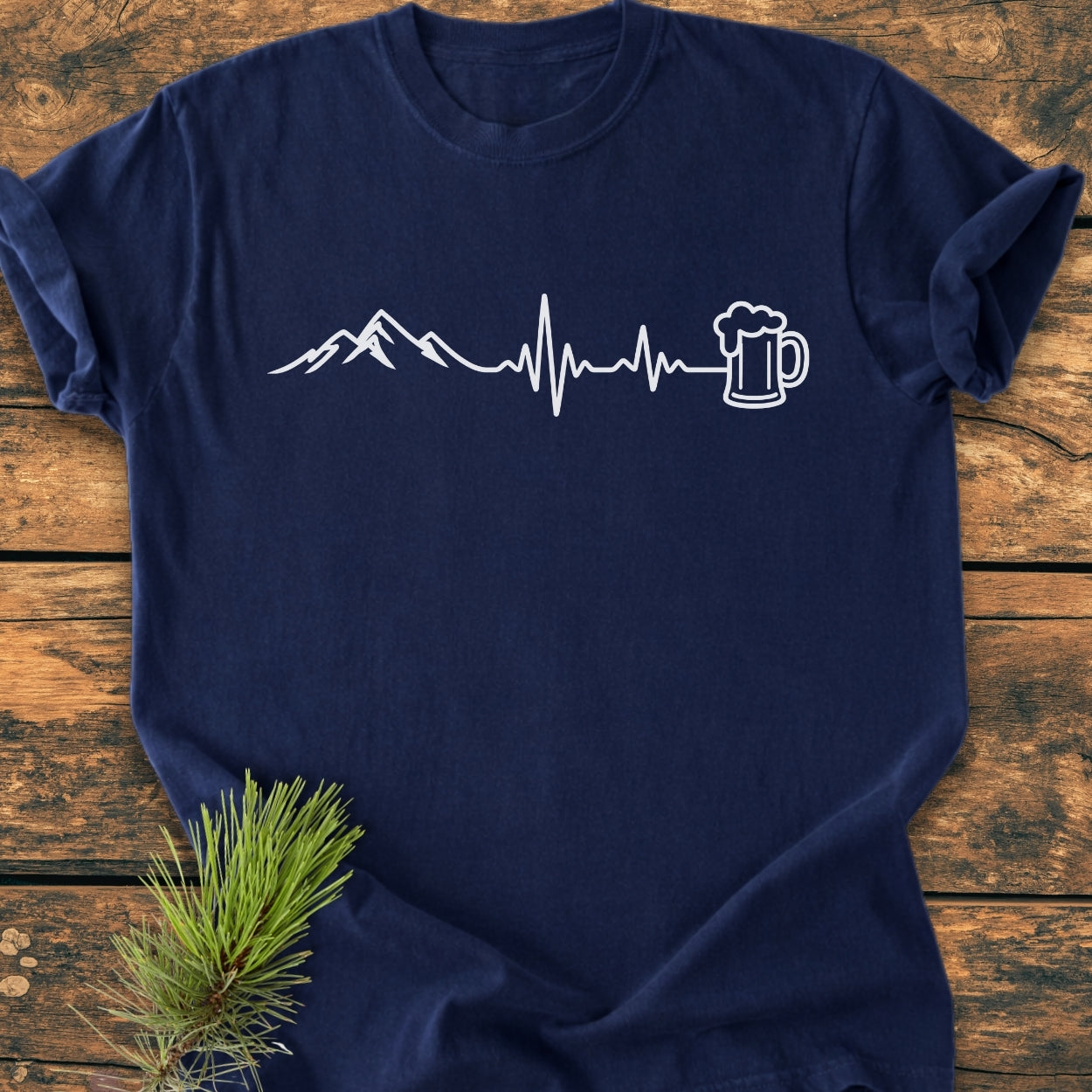 Mountains And Beer - Vintage Wash Tee features a white mountain and beer mug graphic on a soft navy-blue T-shirt, laid on wood with pine. Perfectly combines comfort, durability, and style.