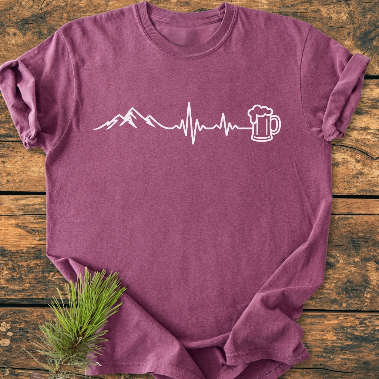 Mountains And Beer - Vintage Wash Tee featuring a white mountain and heartbeat line design with a beer mug laid on rustic wood, enhanced by a pine sprig for a cozy, durable look.