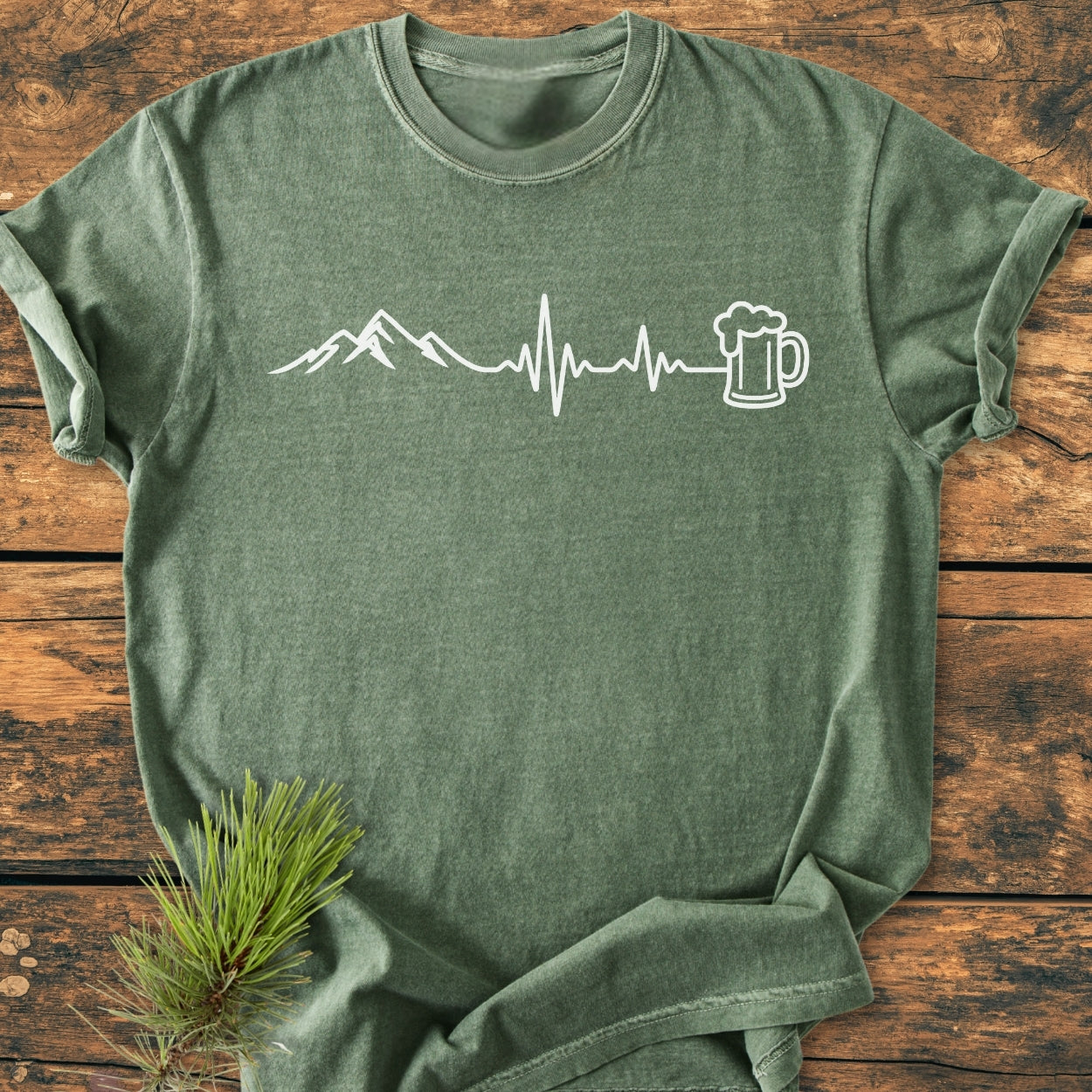 Mountains And Beer - Vintage Wash Tee: Olive-green T-shirt with mountain and EKG-beer graphic, laid on wood. Features a relaxed fit and durable stitching, accentuated by a pine branch detail.
