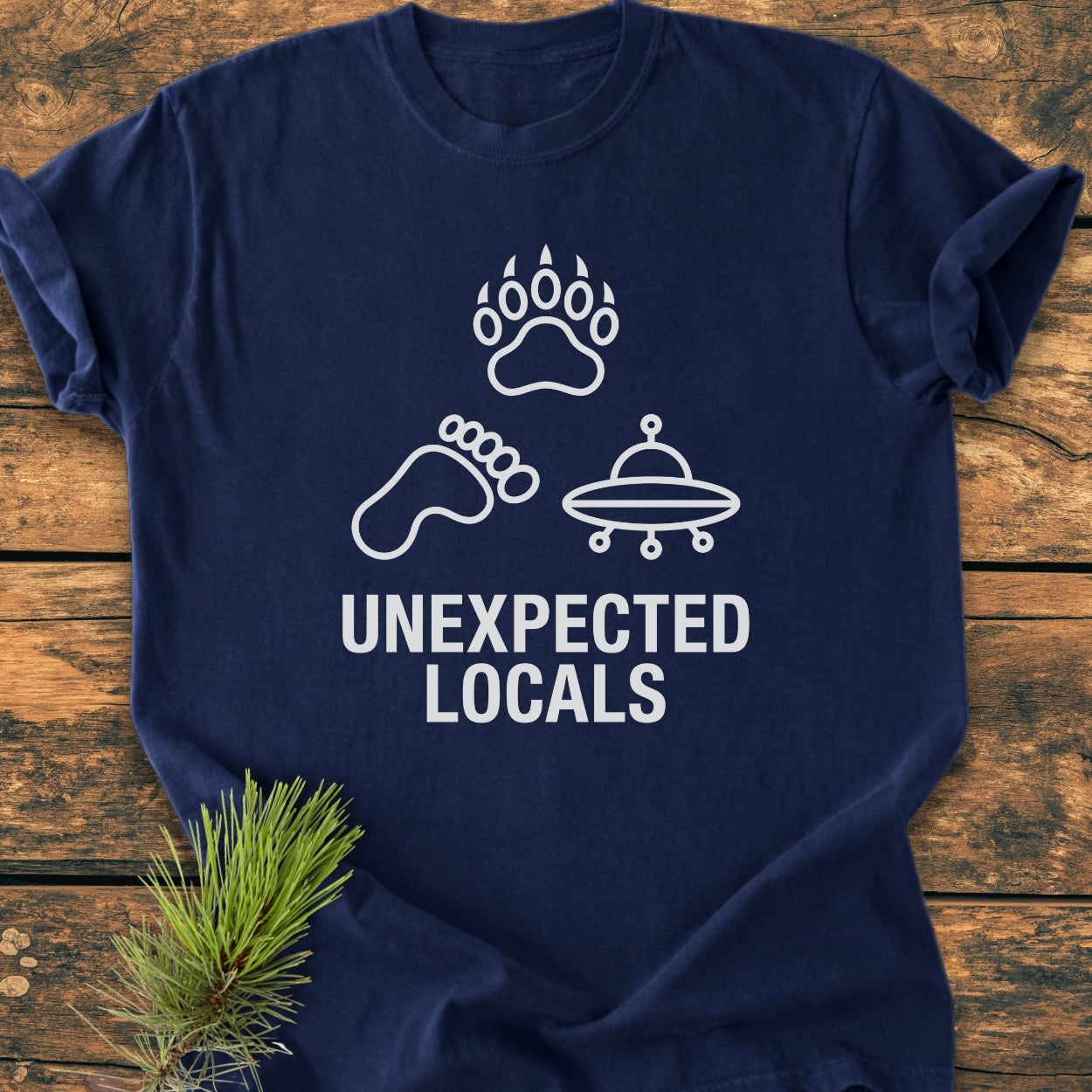 Unexpected Locals - Vintage Wash Tee features navy-blue fabric with white line drawings: a bear paw, Bigfoot footprint, and UFO, arranged in a triangle, with bold UNEXPECTED LOCALS text beneath.
