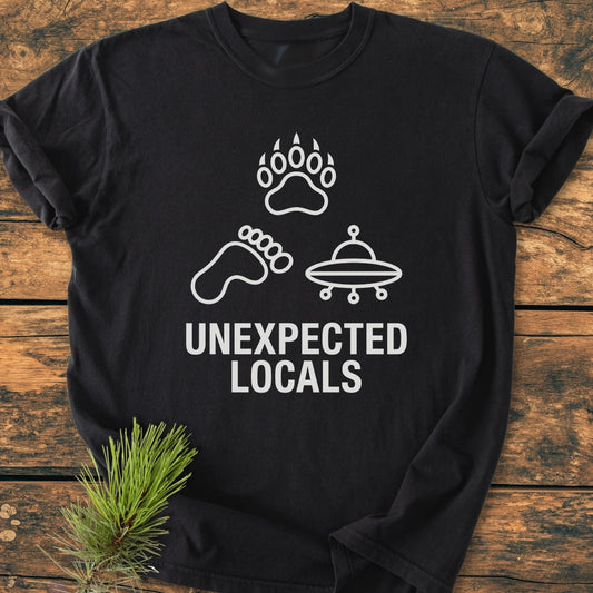 Unexpected Locals - Vintage Wash Tee features a black T-shirt with white line-art icons: bear paw, Bigfoot footprint, UFO. Caption reads 'UNEXPECTED LOCALS.' laid on wooden surface.