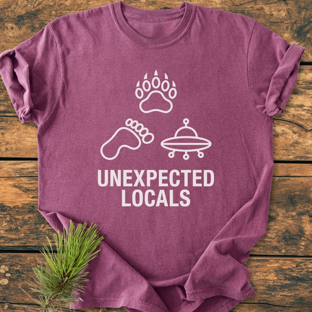 Unexpected Locals - Vintage Wash Tee featuring white icons of a bear paw, Bigfoot footprint, and flying saucer on heathered plum, with pine needles beside it, showcasing a relaxed fit and cozy fabric.