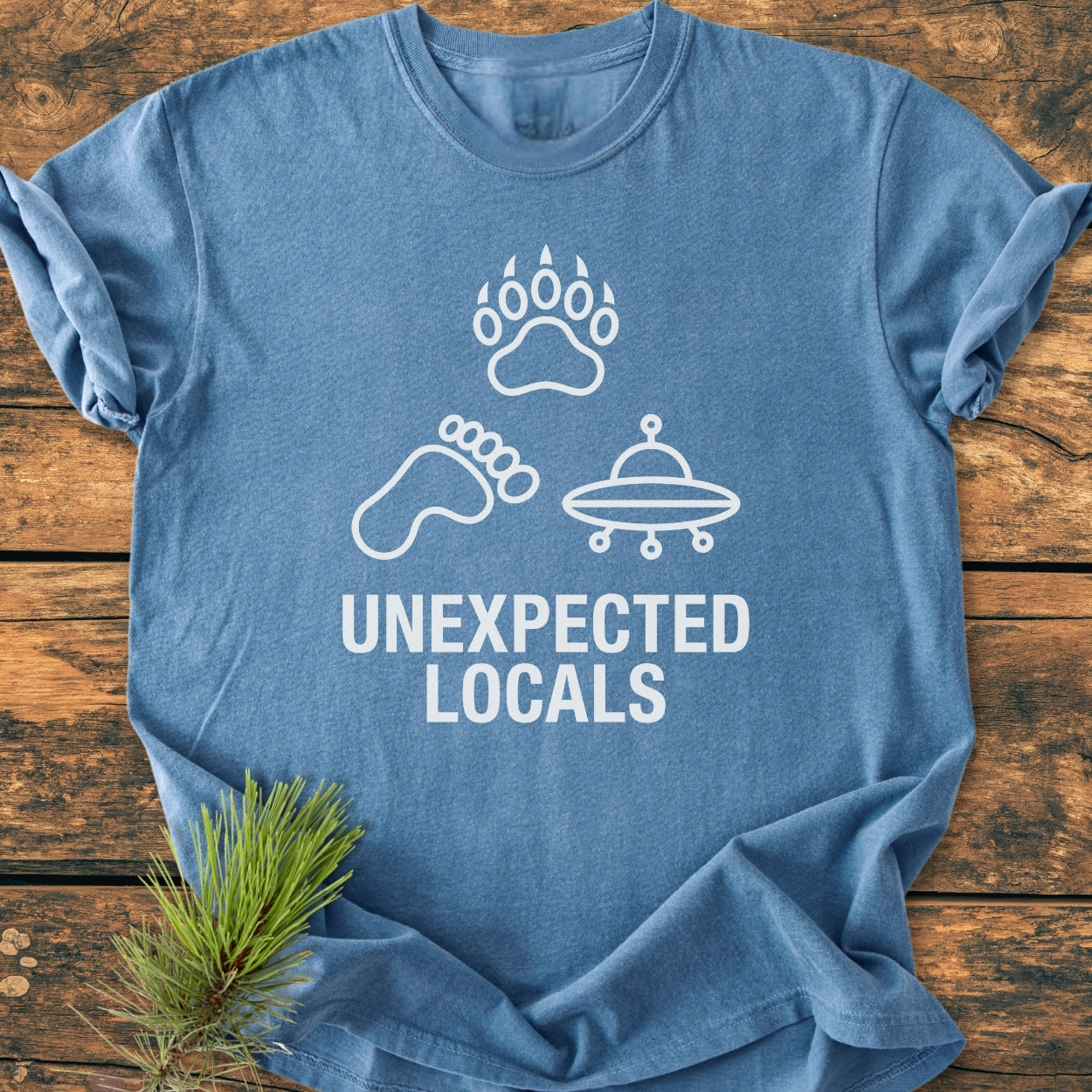 Unexpected Locals - Vintage Wash Tee features blue fabric, white icons of a bear paw, Bigfoot footprint, and UFO above “UNEXPECTED LOCALS,” with a pine sprig beside it on a wooden surface.