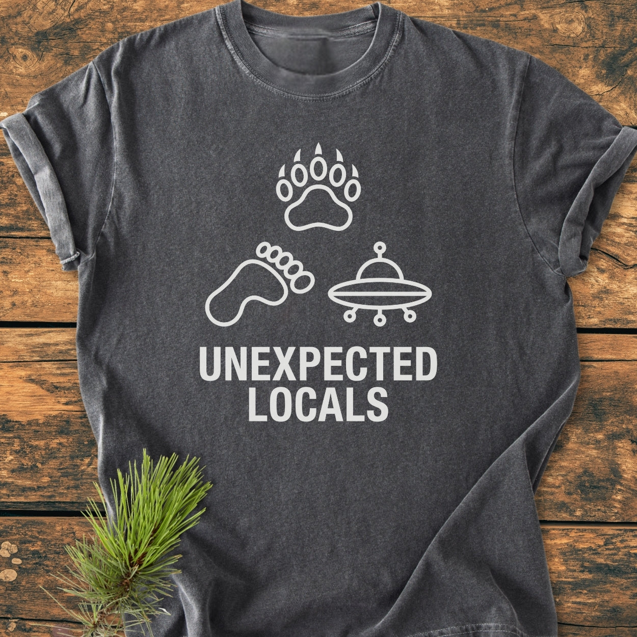 Unexpected Locals - Vintage Wash Tee featuring a charcoal-gray color, displays a bear paw, Bigfoot footprint, and UFO icons with the slogan UNEXPECTED LOCALS. Laid on wood with a pine sprig accent.