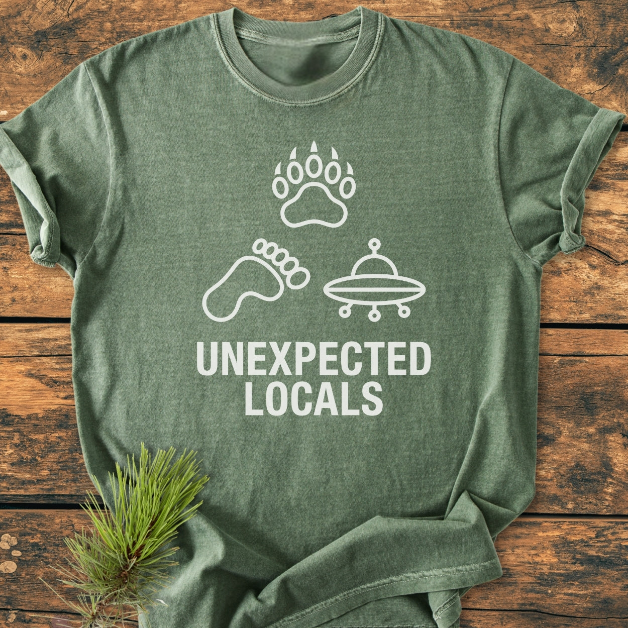 Unexpected Locals - Vintage Wash Tee features a heather-green color with a white graphic of a bear paw, Bigfoot print, and UFO, plus bold text, laid on wood with a pine sprig.