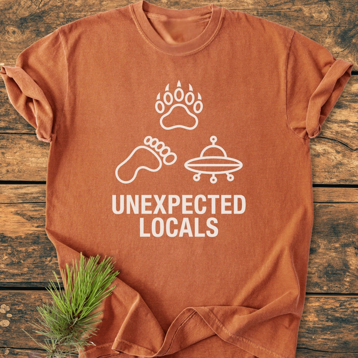 Unexpected Locals - Vintage Wash Tee featuring a rust-orange color with white line-icons of a bear paw, Bigfoot foot, and UFO, plus bold text. Laid on wood with a pine sprig accent.