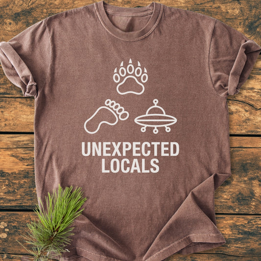Unexpected Locals - Vintage Wash Tee featuring a bear paw, human footprint, and UFO icons above bold text, laid on wood with pine sprig. Soft, durable garment-dyed cotton, relaxed fit.