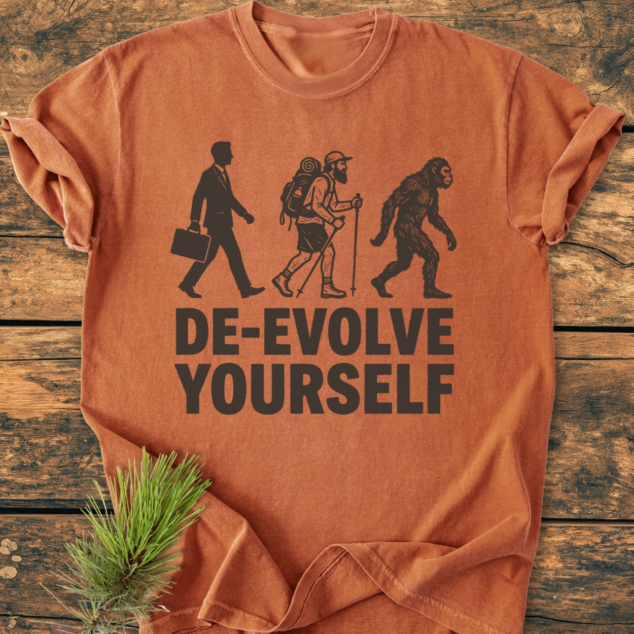 Rust-orange De-Evolve Yourself - Vintage Wash Tee featuring three black silhouettes: businessman, hiker, and ape, with bold text. Laid on wooden surface, showcasing its relaxed fit and durable stitching.