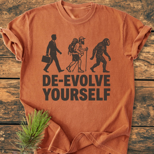 Rust-orange De-Evolve Yourself - Vintage Wash Tee featuring three black silhouettes: businessman, hiker, and ape, with bold text. Laid on wooden surface, showcasing its relaxed fit and durable stitching.