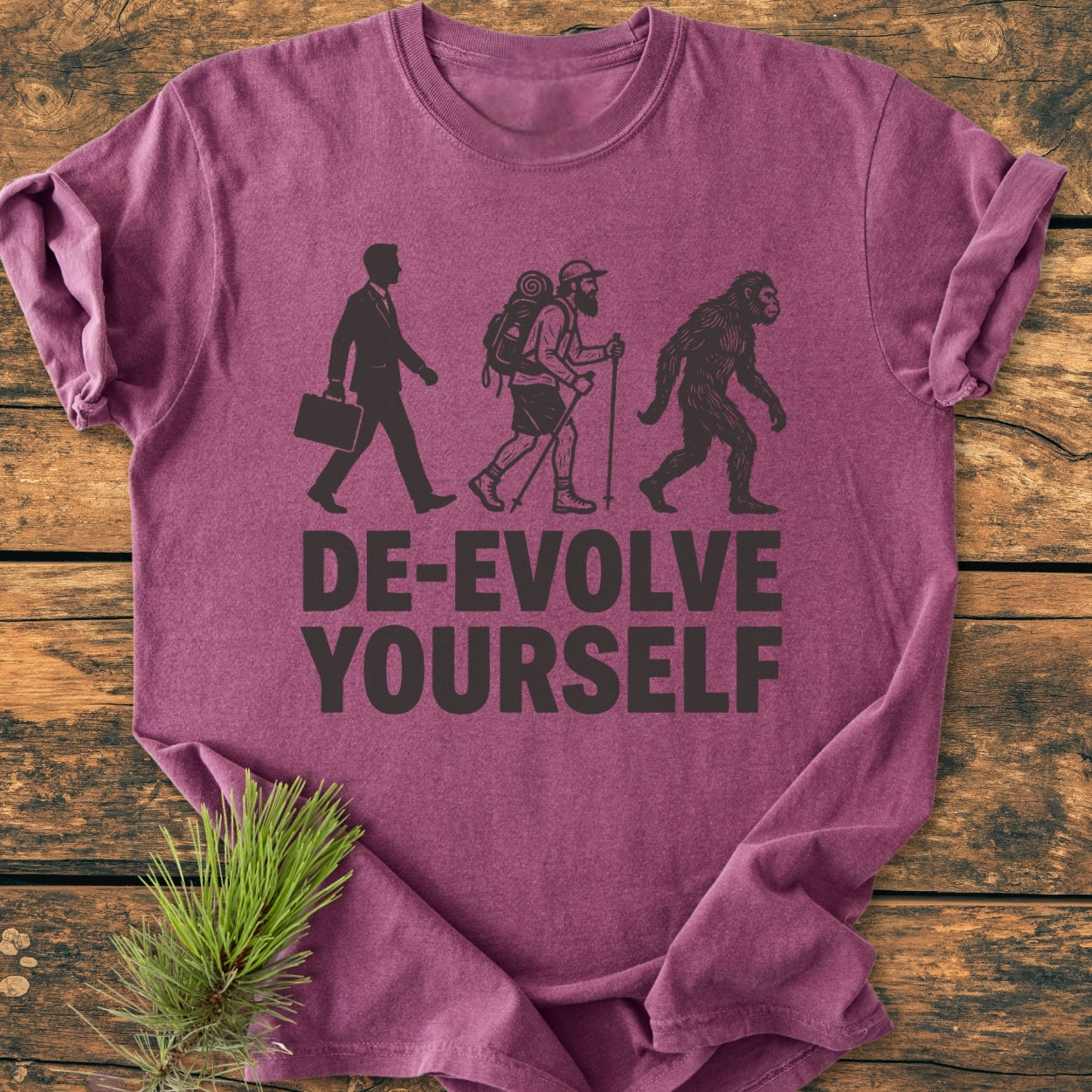 De-Evolve Yourself - Vintage Wash Tee, a heather-purple T-shirt featuring a “reverse evolution” graphic, laid on wood. Includes a business suit man, hiker, and ape with slogan.
