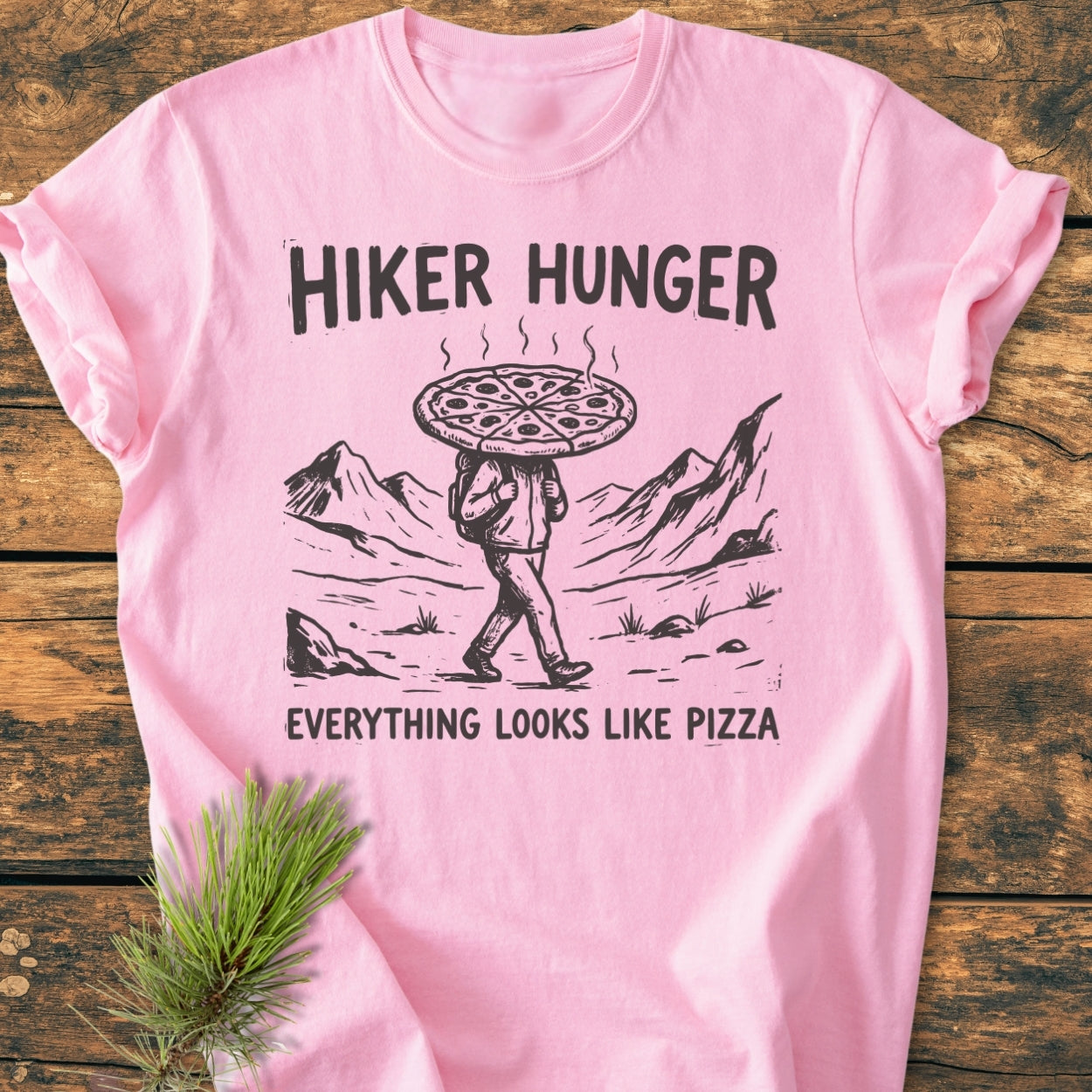 Hiker Hunger - Vintage Wash Tee featuring a black-ink hiker with pizza head graphic on light-pink fabric, laid on wood with pine sprig, emphasizing comfort and durability.
