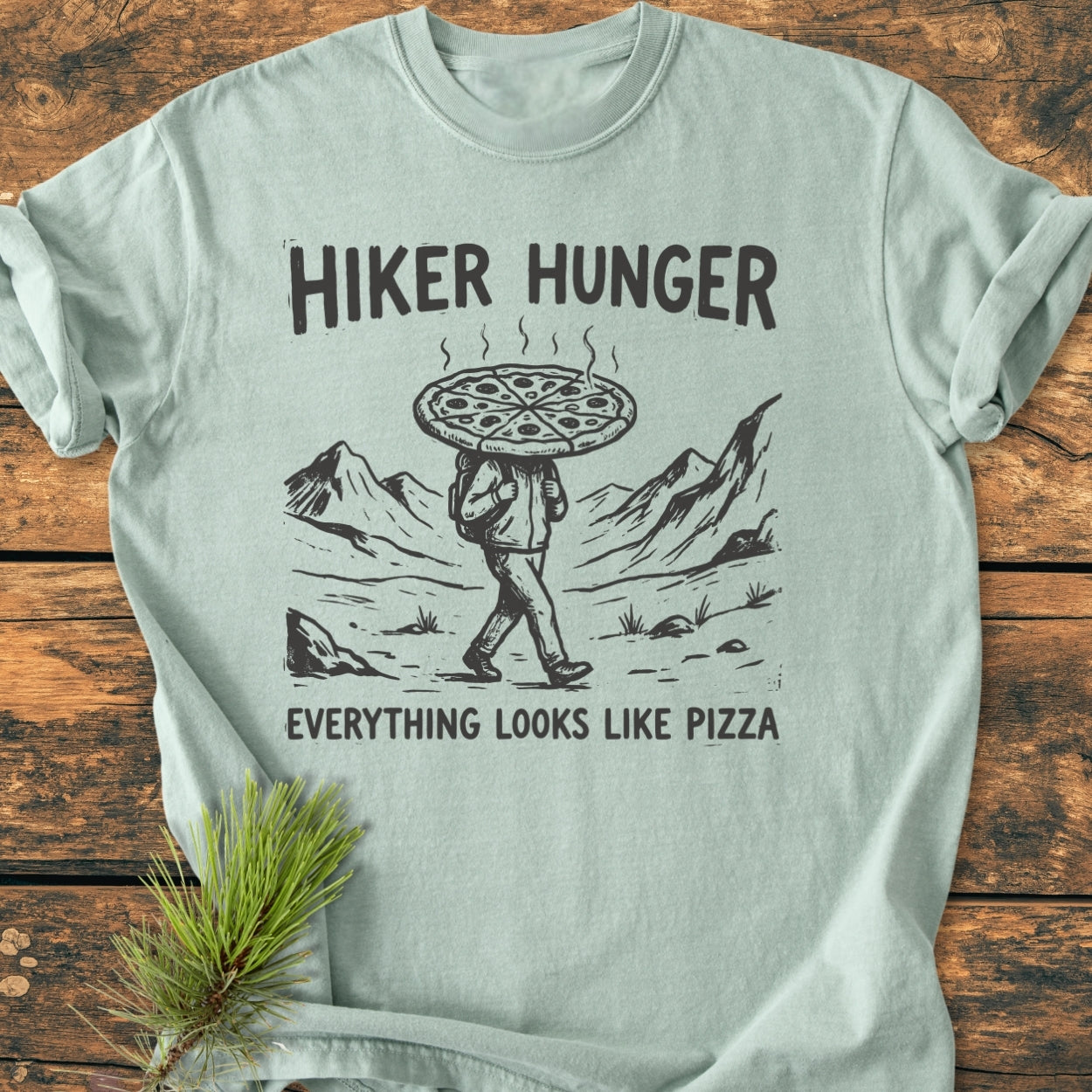 Hiker Hunger - Vintage Wash Tee: Pale-green T-shirt featuring a black line-drawing of a hiker with a pizza pie head, surrounded by mountains. Text reads “HIKER HUNGER” above and “EVERYTHING LOOKS LIKE PIZZA” below.
