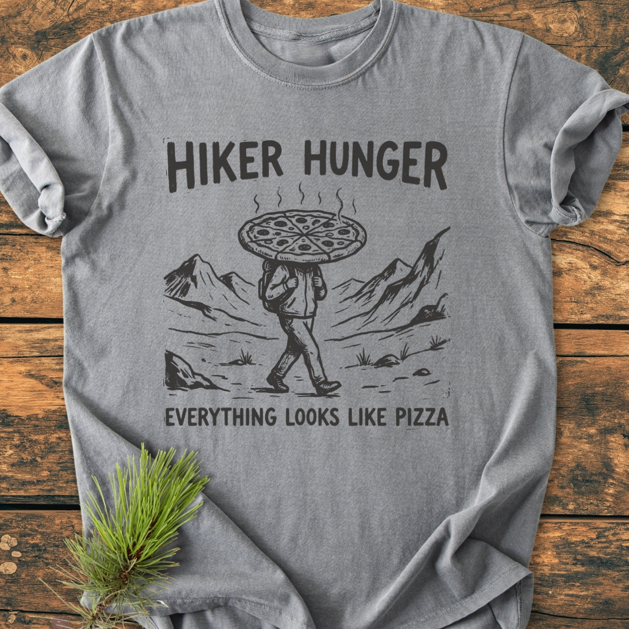 Hiker Hunger - Vintage Wash Tee featuring a hand-drawn hiker with a pizza pie head, set against mountains, laid on wood with a pine sprig; made from soft, durable, 100% ring-spun cotton.
