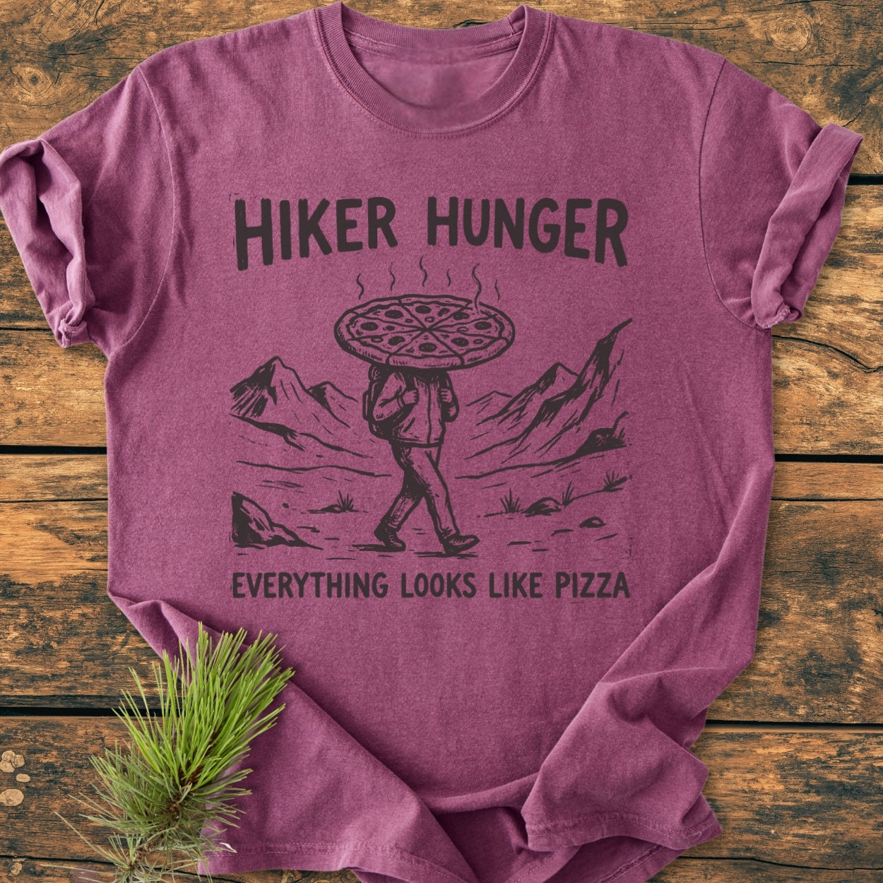 Hiker Hunger - Vintage Wash Tee featuring a unique design of a pizza-headed hiker amidst mountains, laid on wood with a pine sprig, showcasing its relaxed fit and garment-dyed fabric.