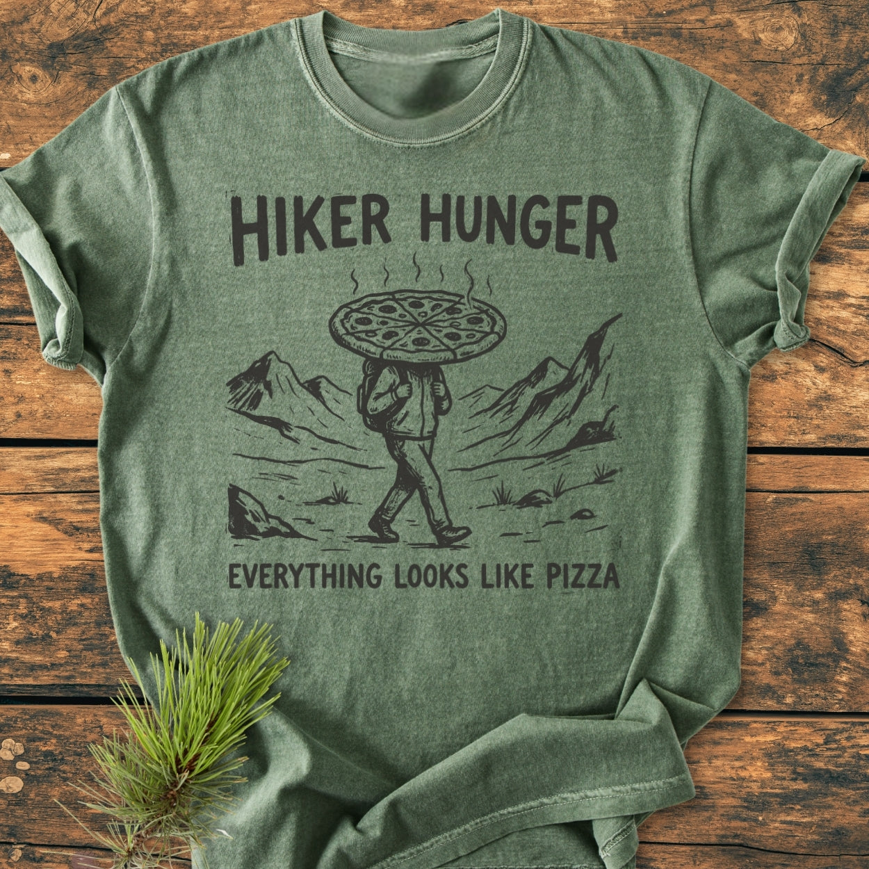 Hiker Hunger - Vintage Wash Tee features a black illustration of a hiker with a pizza head on heather-green fabric, laid on rustic wood with a pine branch, exuding cozy, relaxed vibes.