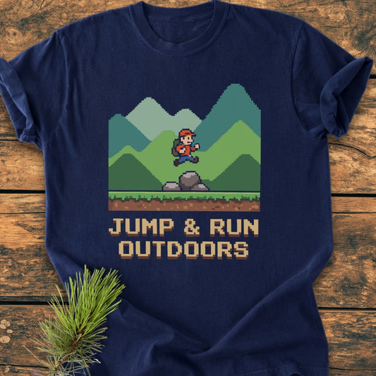 Jump And Run Outdoors - Vintage Wash Tee featuring an 8-bit hiker graphic over mountains, laid flat on wood. Accompanied by a pine sprig, the navy-blue tee embodies a cozy, relaxed fit.