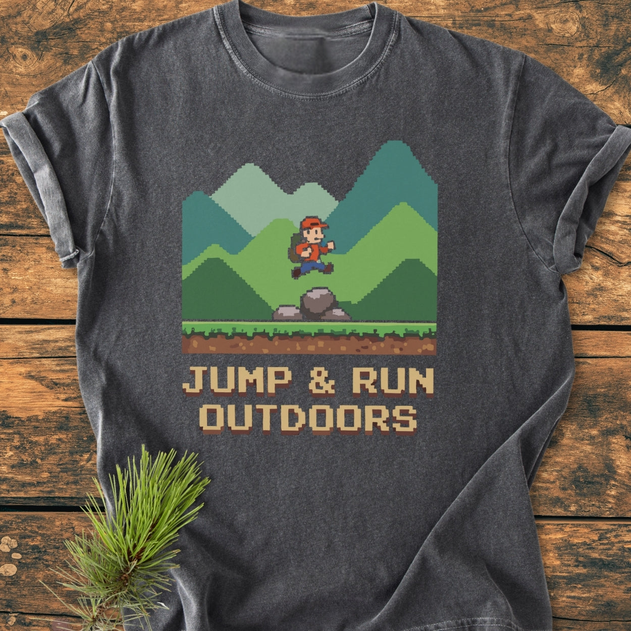 Jump And Run Outdoors - Vintage Wash Tee features a pixel-art hiker jumping over a rock with green mountains. Laid on rustic wood, the tee offers a cozy fit with robust double-needle stitching.