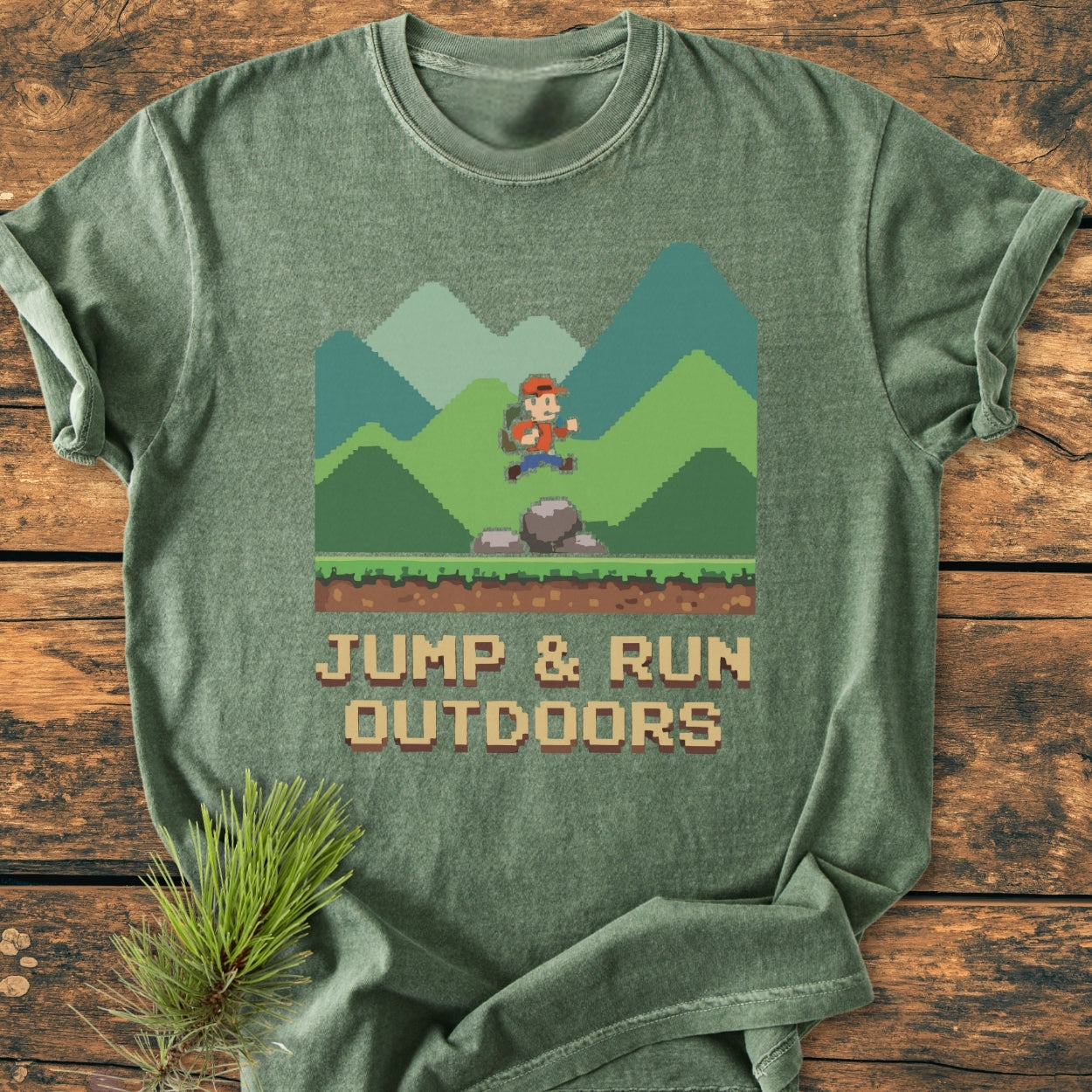 Jump And Run Outdoors - Vintage Wash Tee features a heather-green T-shirt with an 8-bit hiker scene and slogan, laid on wood. Pine needles accent the soft-washed, relaxed-fit design.
