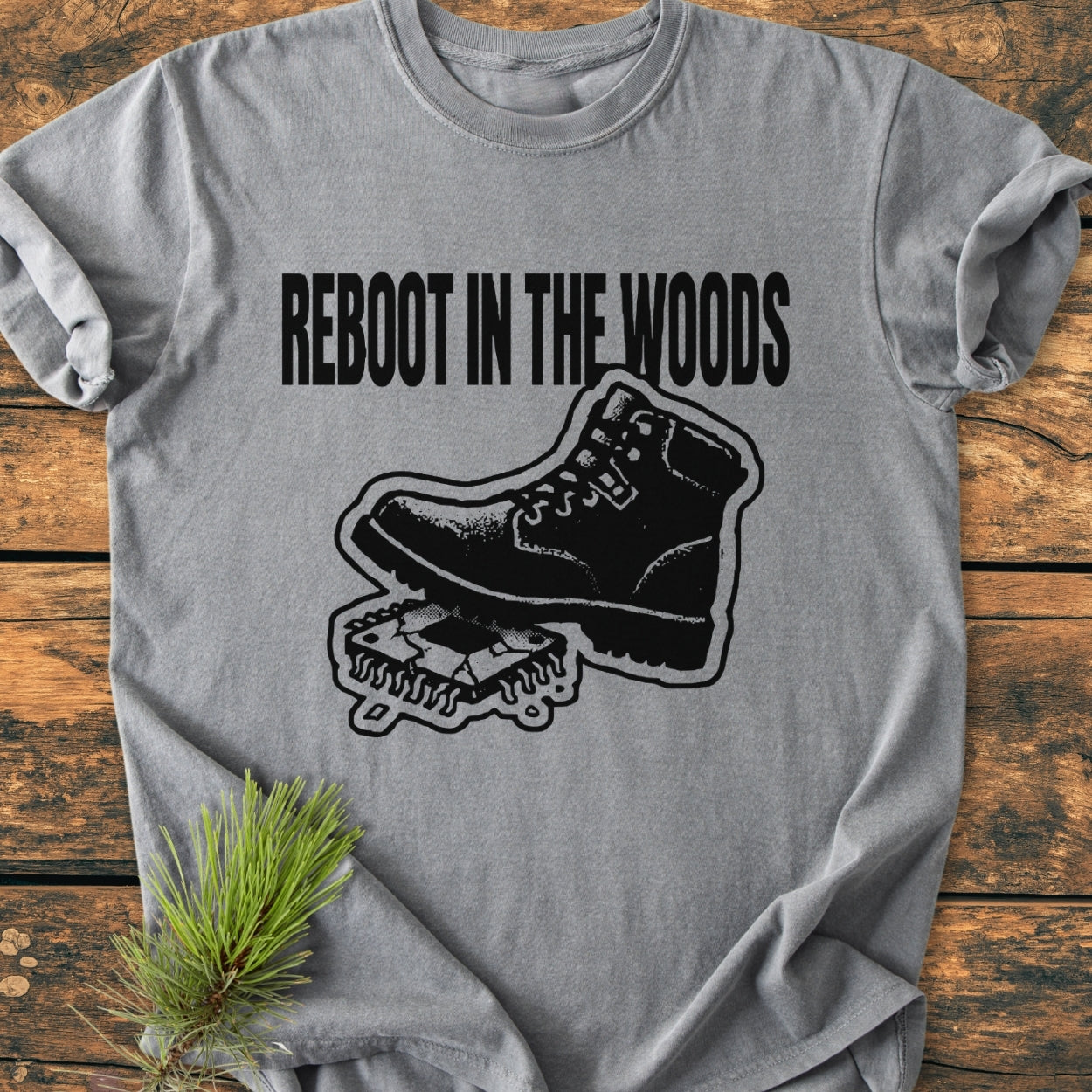 Reboot In The Wood - Vintage Wash Tee featuring a hiking boot graphic rebooting a microchip, laid on a wooden surface with pine needles accentuating the outdoor theme.