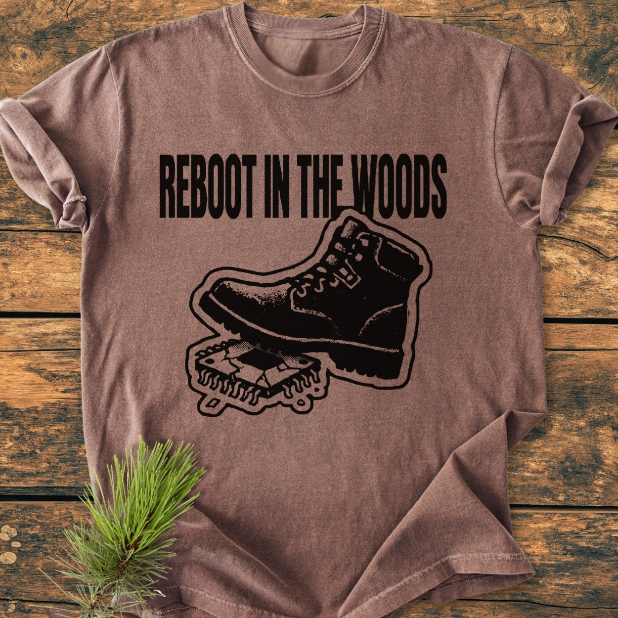 Reboot In The Woods - Vintage Wash Tee displayed on a rustic surface, featuring a hiking boot graphic over a circuit board, symbolizing 'reboot.' Cozy, relaxed fit with double-needle stitching for durability.