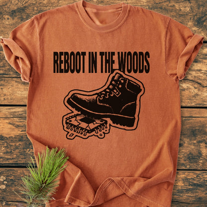 Reboot In The Woods - Vintage Wash Tee: A folded burnt-orange T-shirt featuring a hiking boot graphic crushing a circuit chip with the slogan above, laid on a wooden surface with a pine bough accent.
