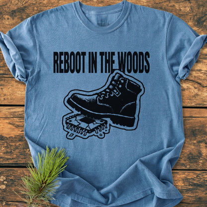 Heather-blue Reboot In The Woods - Vintage Wash Tee with bold black slogan and hiking boot graphic on a circuit board, laid on rustic wood, with a pine sprig beside it.