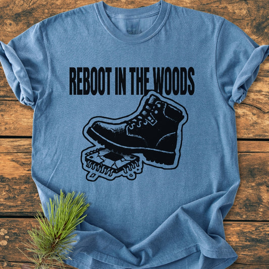 Heather-blue Reboot In The Woods - Vintage Wash Tee with bold black slogan and hiking boot graphic on a circuit board, laid on rustic wood, with a pine sprig beside it.