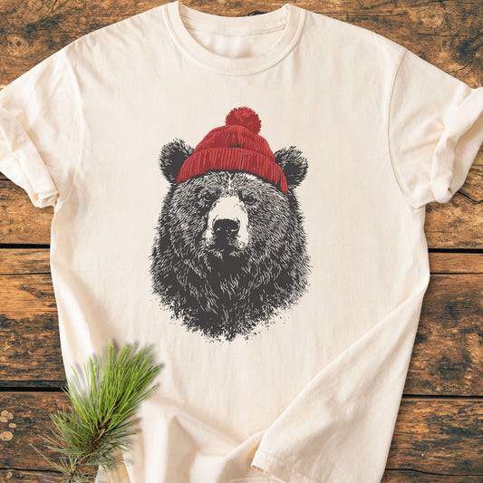 Bear With Beanie - Vintage Wash Tee on wood, featuring a sketched bear in a red winter hat. Made from cozy, soft-washed 100% ring-spun cotton. Pine needles accent the scene.